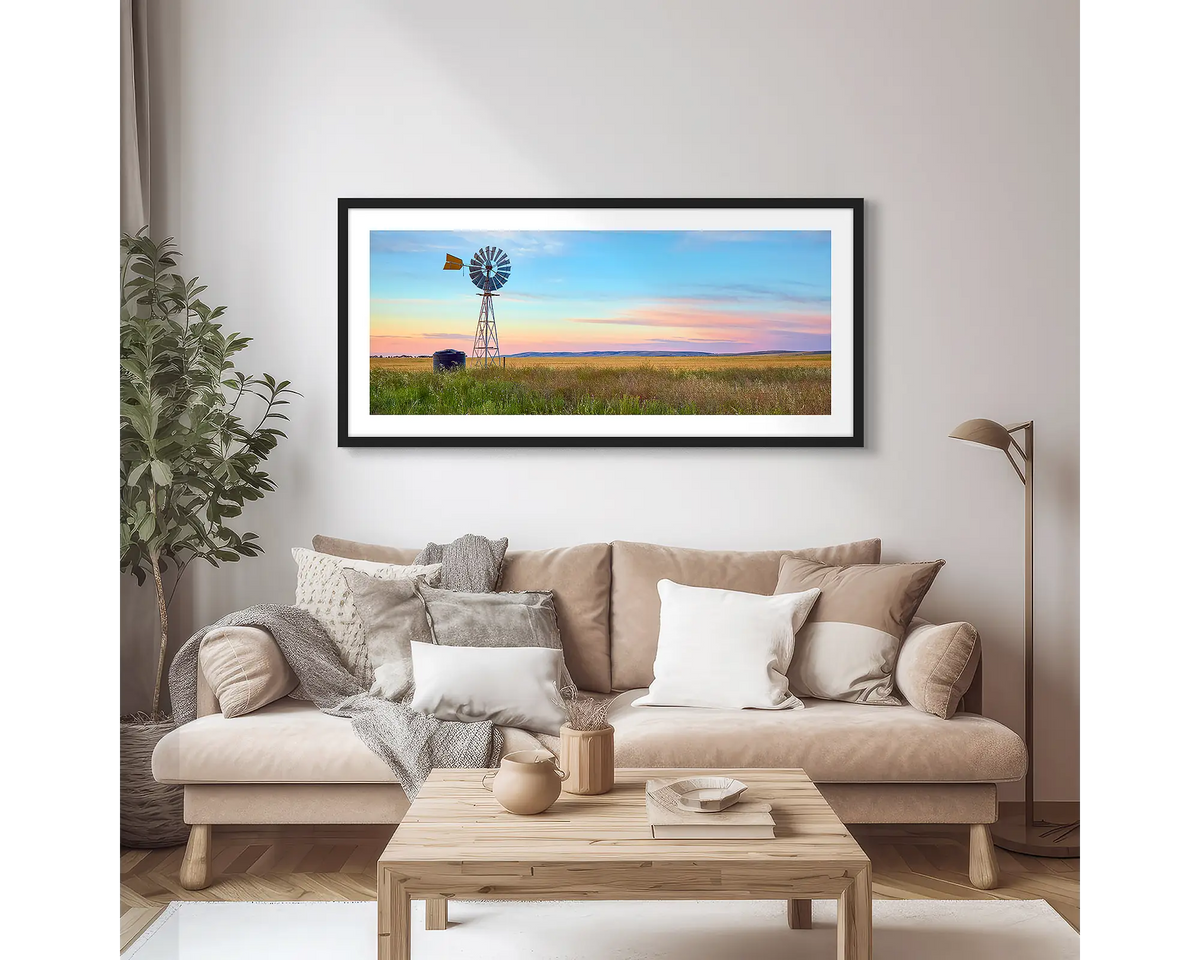 Painted Morning. Wall art print of a windmill on a farm, with a colourful sunrise, with a black frame hung in an apartment living room. 