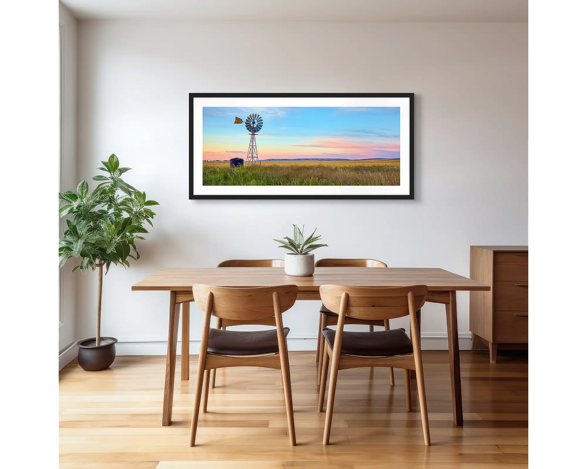 Painted Morning. Wall art print of a windmill on a farm, with a colourful sunrise, with a black frame hung in a dining area. 