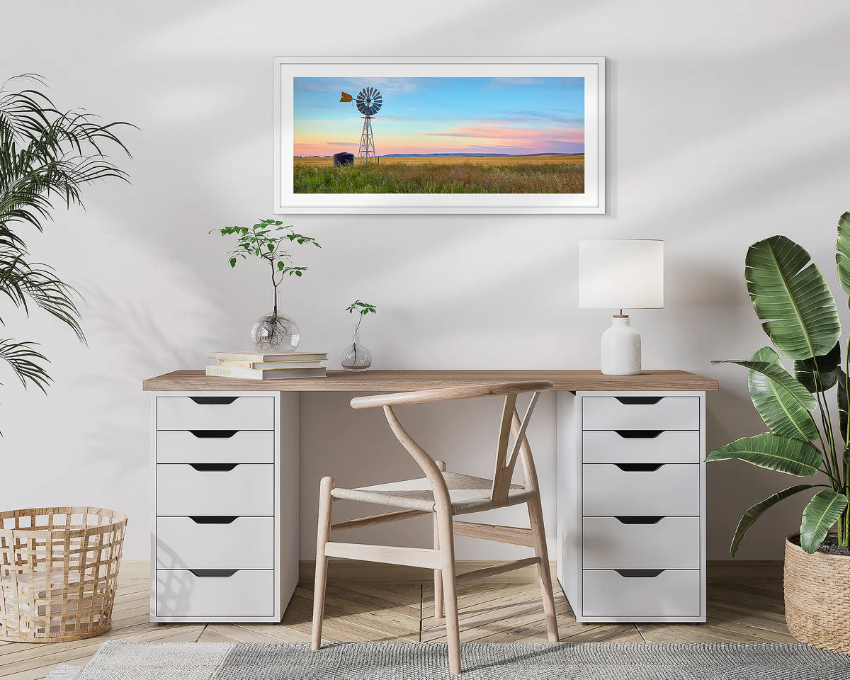 Painted Morning. Wall art print of a windmill on a farm, with a colourful sunrise, with a white frame hung above a small desk. 