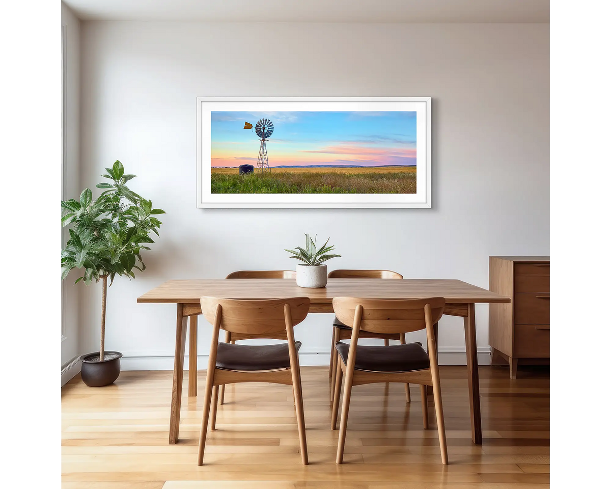 Painted Morning. Wall art print of a windmill on a farm, with a colourful sunrise, with a white frame hung in a dining area. 