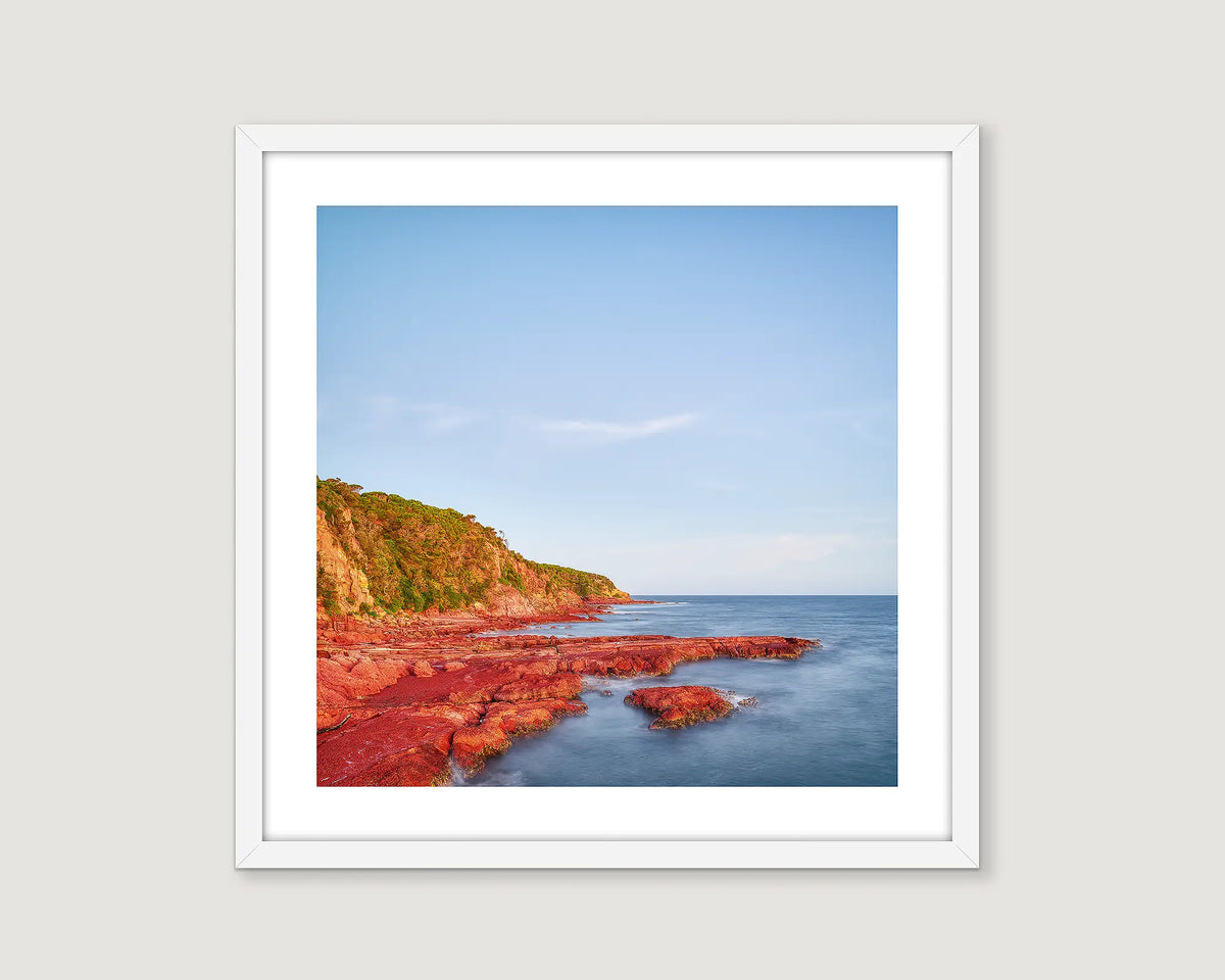 Framed photograph of a red cliff and the ocean with a blue sky.