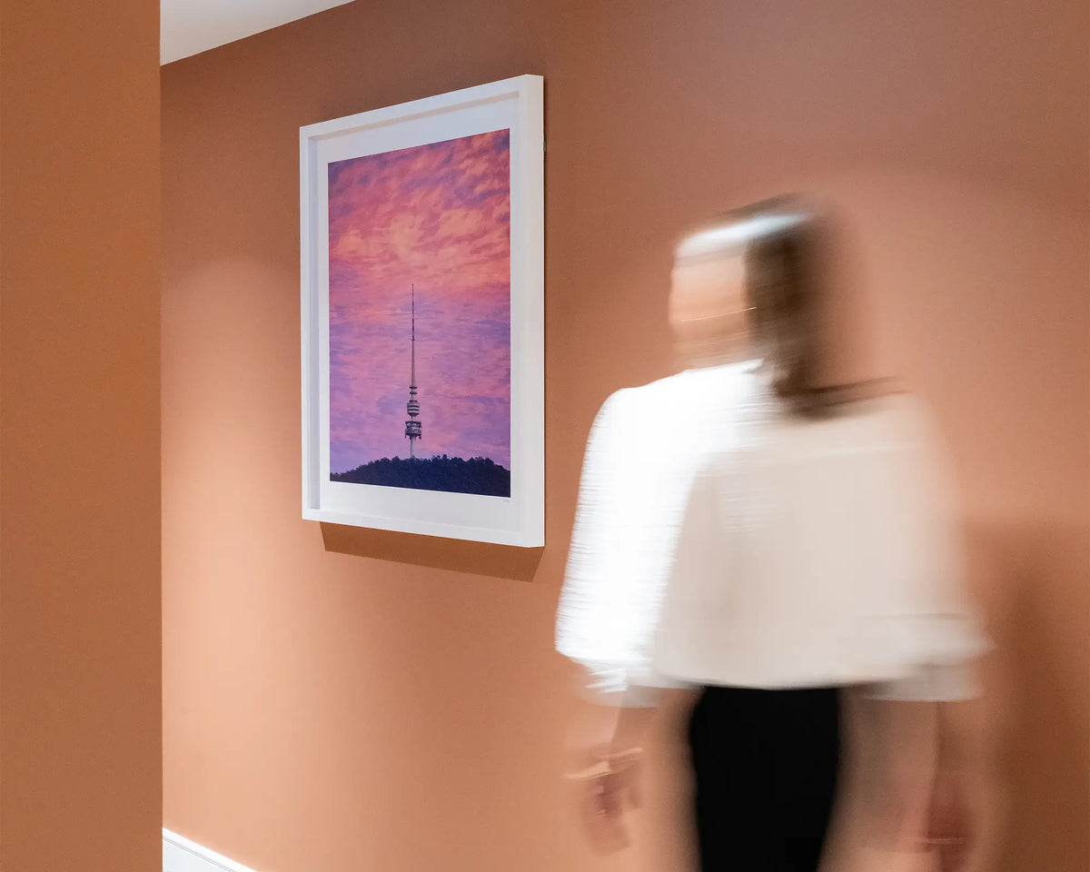 Pointer. Sunrise at Telstra Tower Canberra, with white frame hung on terracotta coloured wall, with person walking toward.