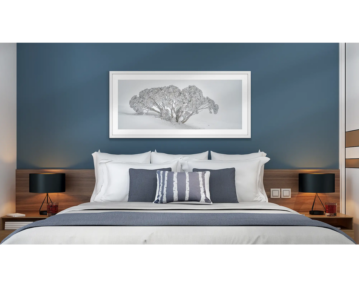 Revealed. Wall art print of a snow gum covered in snow,, framed with a white frame and hung above a bed in a hotel room. 