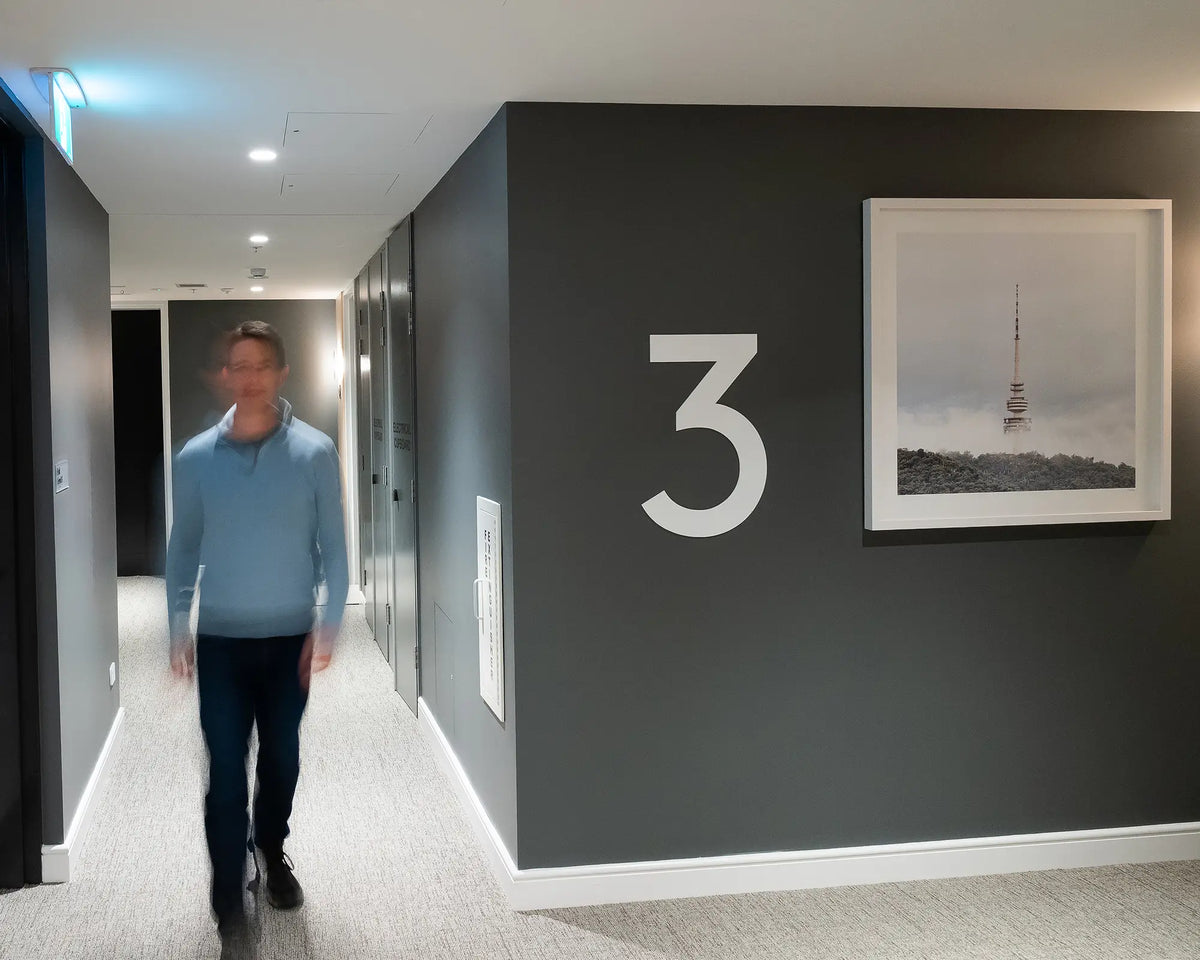 Rising. Black Mountain Tower in fog with white frame, hung on grey wall third floor apartment building, with person walking down corridor.