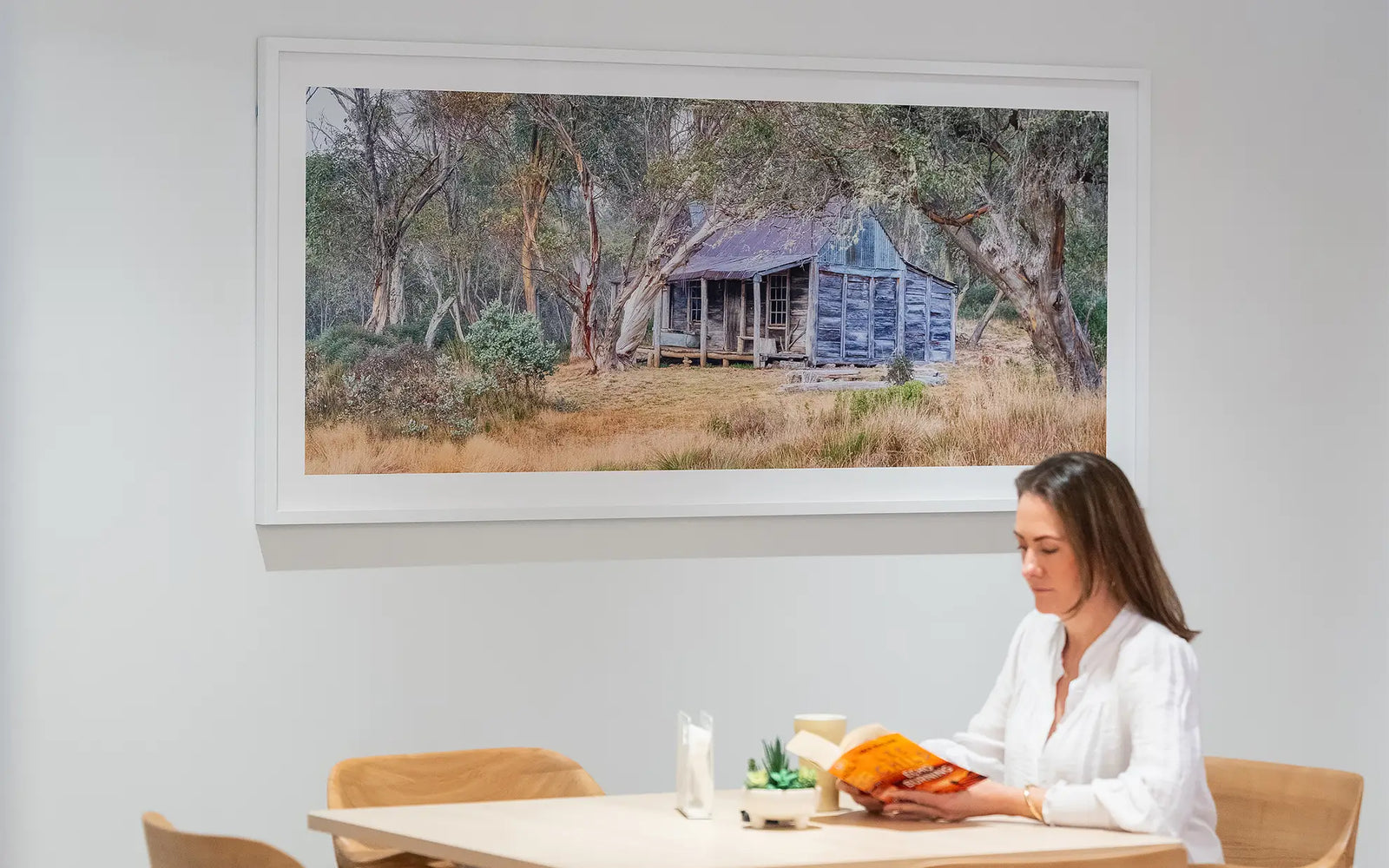 A woman in a white blouse sits at a light wood table reading a book, with a large framed Scott Leggo landscape photograph of anWheelers Hut hanging on the wall beside her.
