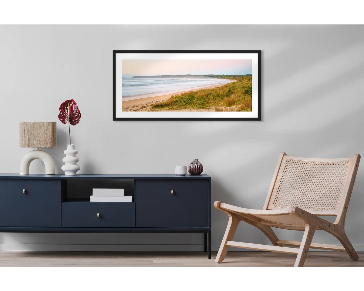 Seaside Dawn. Wall art print of a sunrise at Durras Beach, with a black frame, hung above a side table and chair. 