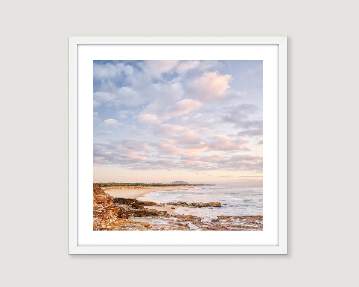 Framed photograph of a beach shoreline with sand and rocks and a blue sky with some clouds.