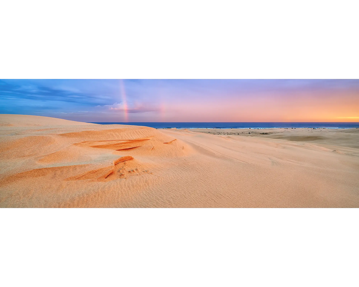 Sand dunes and a rainbow in the sky at sunset.