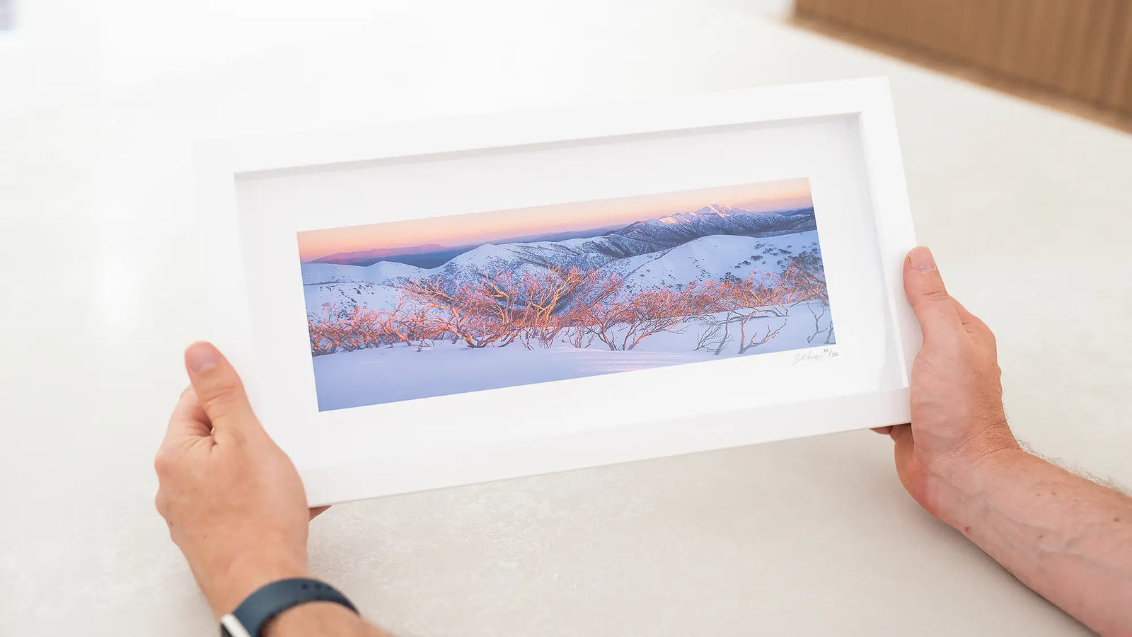 Person holding a framed picture of a snowy sunrise landscape.