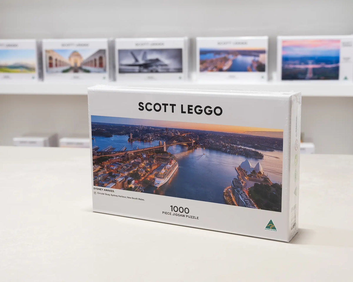 Sydney Awakes 1000 piece jigsaw puzzle box sitting on table in front of other puzzle boxes.