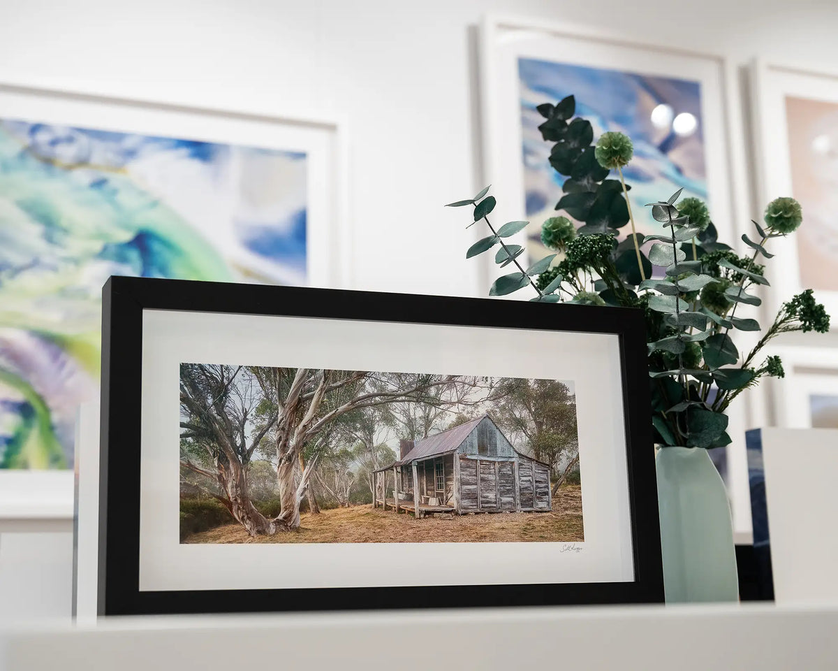 Tales Of The Stockman. Wheelers Hut, Kosciuszko National Park, with black frame, placed on gallery desk with flower arrangement.