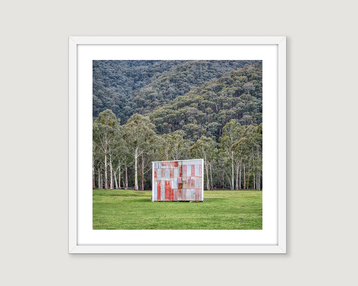 Framed artwork of a red and grey farm shed in a grassy field with trees and mountains in the background.