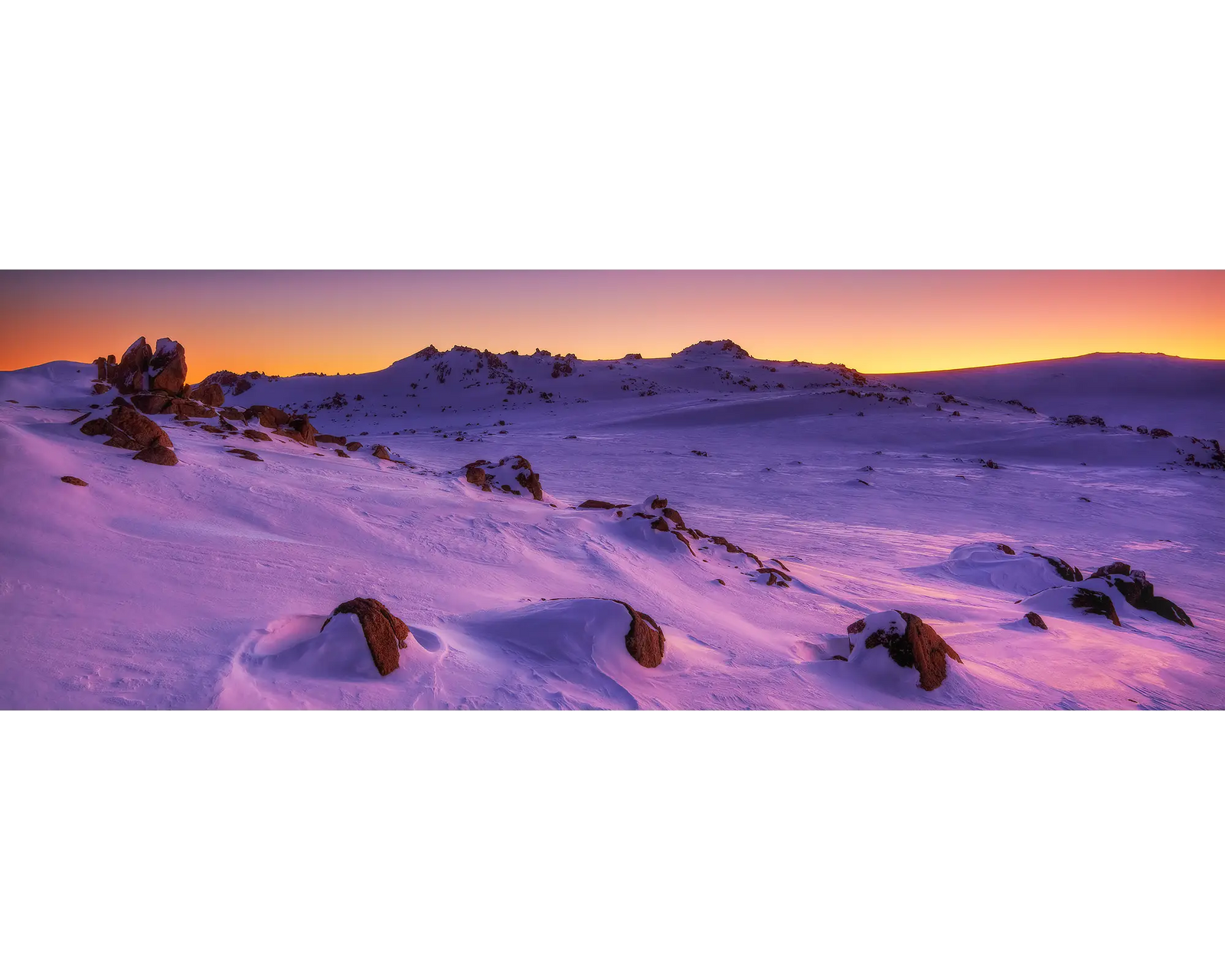 Snowy landscape with rocks at sunset, featuring a pink and purple sky.