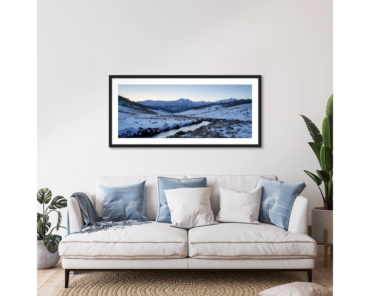 Thredbo Colours. Wall art print with a black frame hung above a couch, featuring the Thredbo River with winter snow.
