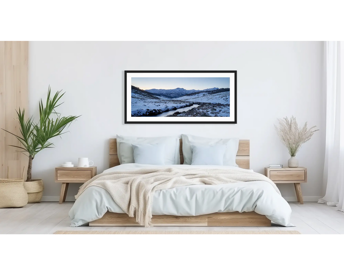Thredbo Colours. Wall art print with a black frame hung above a bed, featuring the Thredbo River with winter snow.