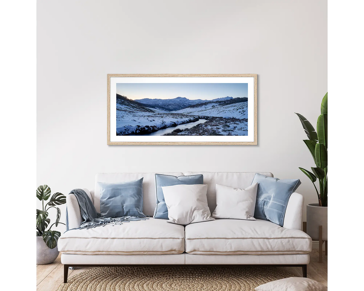 Thredbo Colours. Wall art print with a timber frame hung above a couch, featuring the Thredbo River with winter snow.