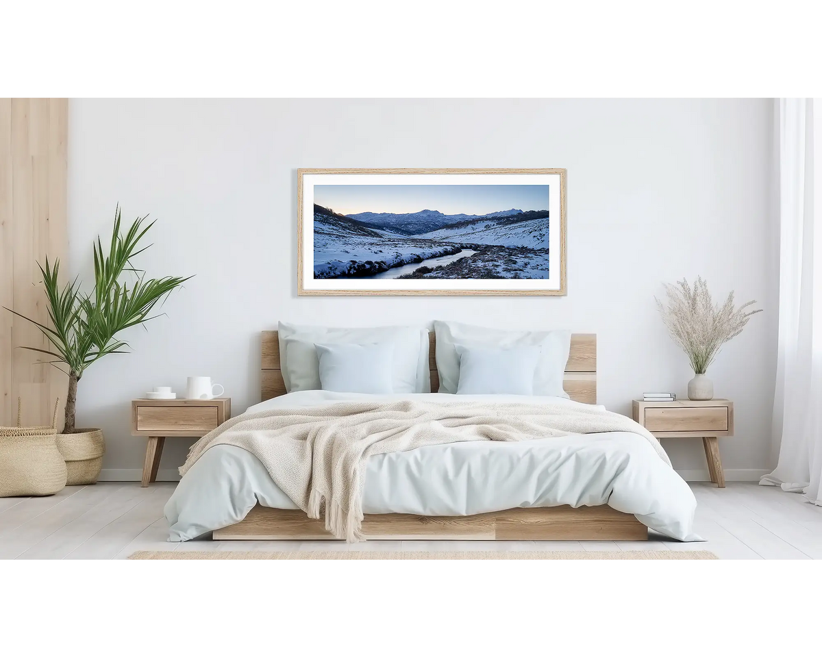 Thredbo Colours. Wall art print with a timber frame hung above a bed, featuring the Thredbo River with winter snow.
