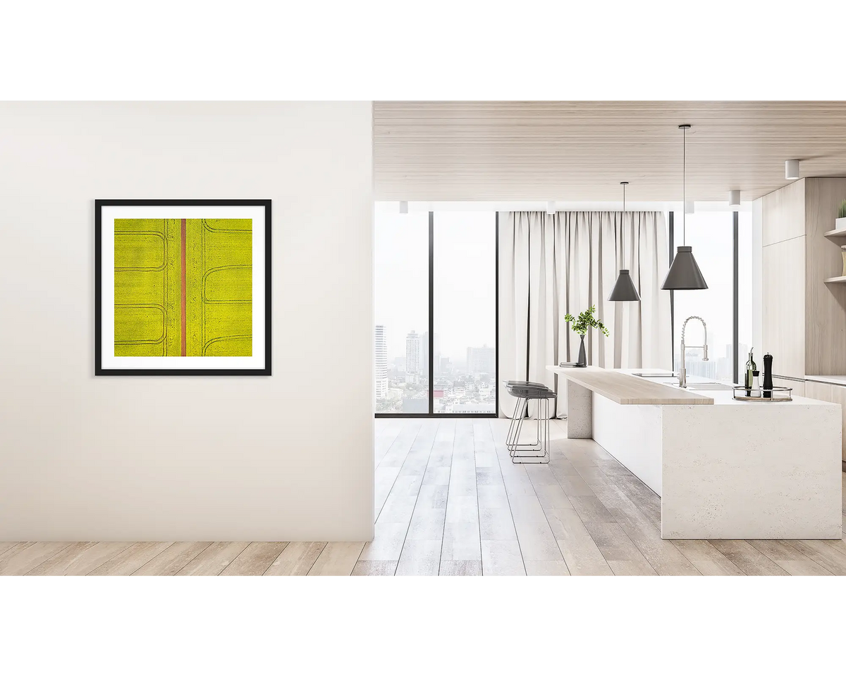 Tracks. Framed wall art print of Canola fields viewed from above, with a black frame, hung on a hallway wall. 