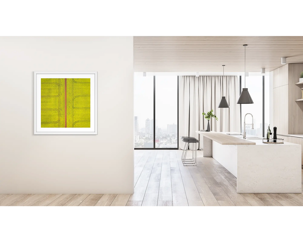 Tracks. Framed wall art print of Canola fields viewed from above, with a white frame, hung on a hallway wall. 