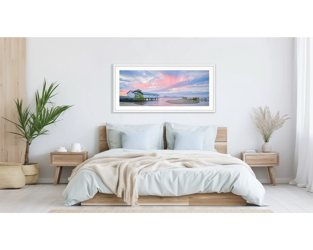 Twilight Blush. Wall art print of a pink sunset over Port Douglas, framed in a white frame and hung above a bed. 
