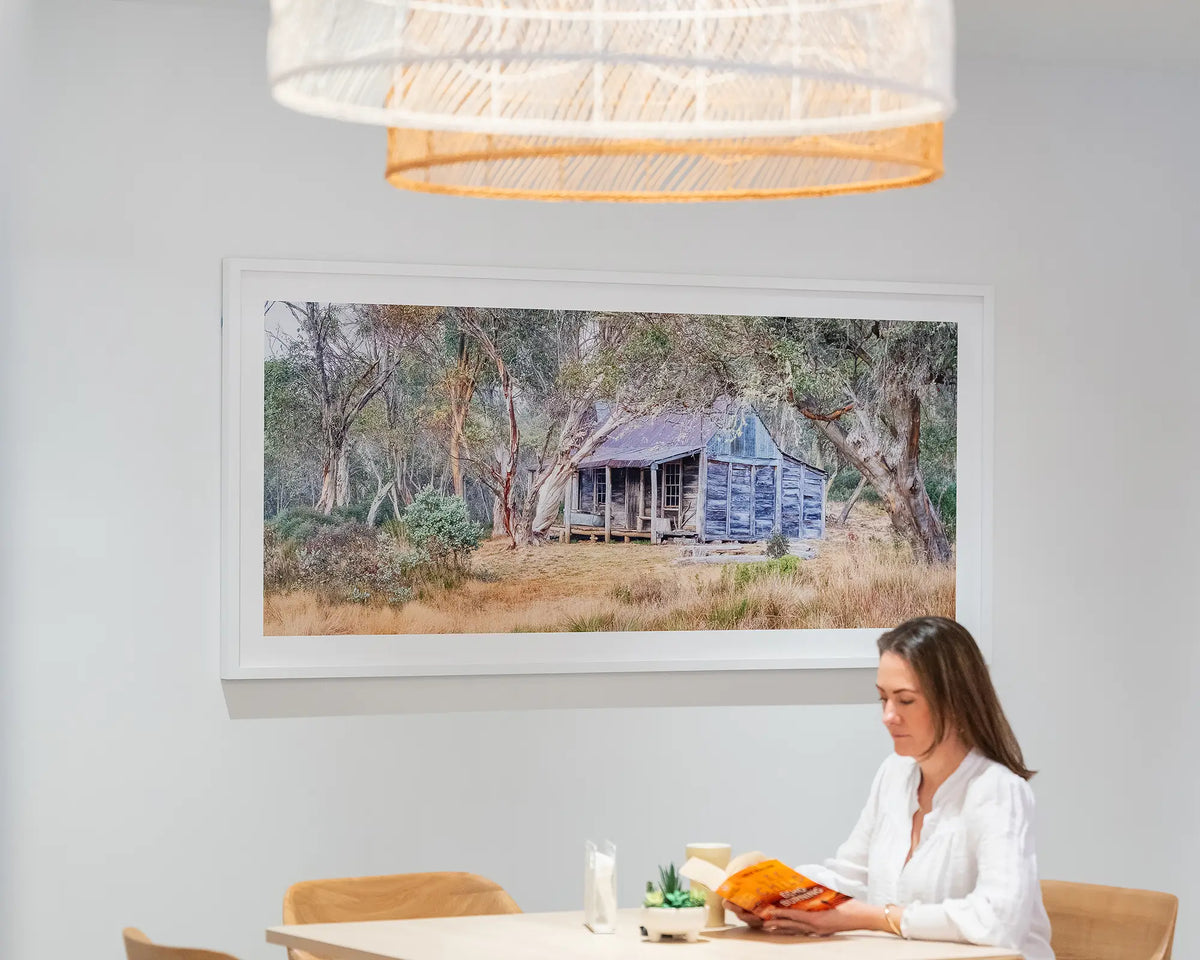 Wheelers Legacy. Wheelers Hut, Kosciuszko National Park, with white frame, hung in dining room with person reading in foreground.